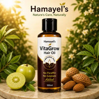 A bottle of Hamayel's VitaGrow Hair Oil stands on a marble surface surrounded by green leaves, a green fruit, and almonds, with a warm blurred floral background. The brand name 'Hamayel's' is displayed at the top of the image.